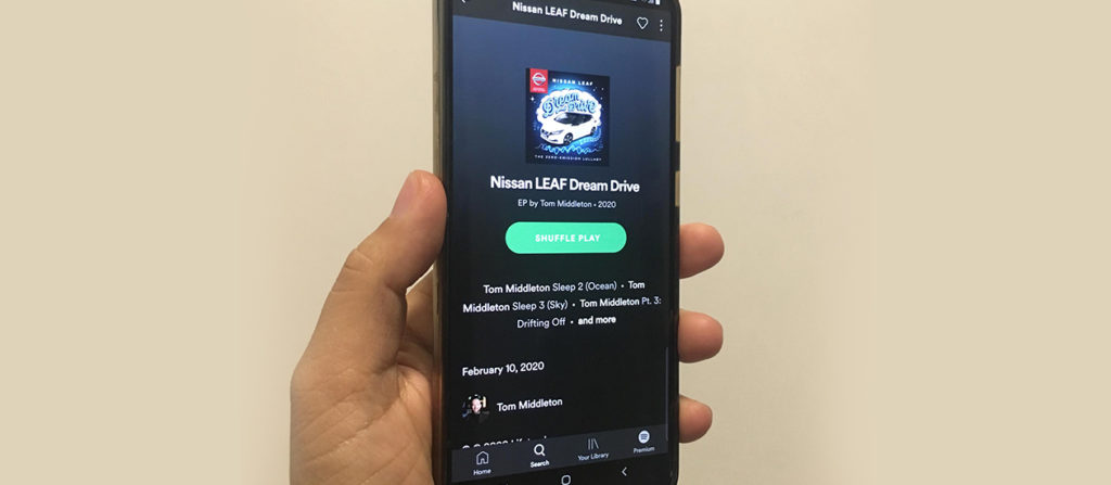 Hand holding up a phone that displays a music app playing Nissan Dream Drive's playlist